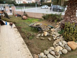 Paysagiste taillant la haie dans un jardin en rénovation. Une brouette de galets et des outils sont disposés sur la pelouse près d'une bordure rocheuse.