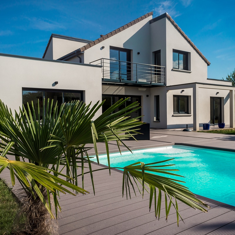 Piscine et terrasse design : l'extérieur d'une maison moderne. Architecture moderne avec façade claire, balcon, et baies vitrées. Piscine turquoise, terrasse grise et grand palmier ensoleillé.