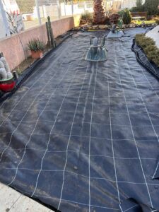 Jardin préparé avec toile de paillage noire à quadrillage blanc, fixée au sol. Brouette renversée et plantes grasses en arrière-plan.