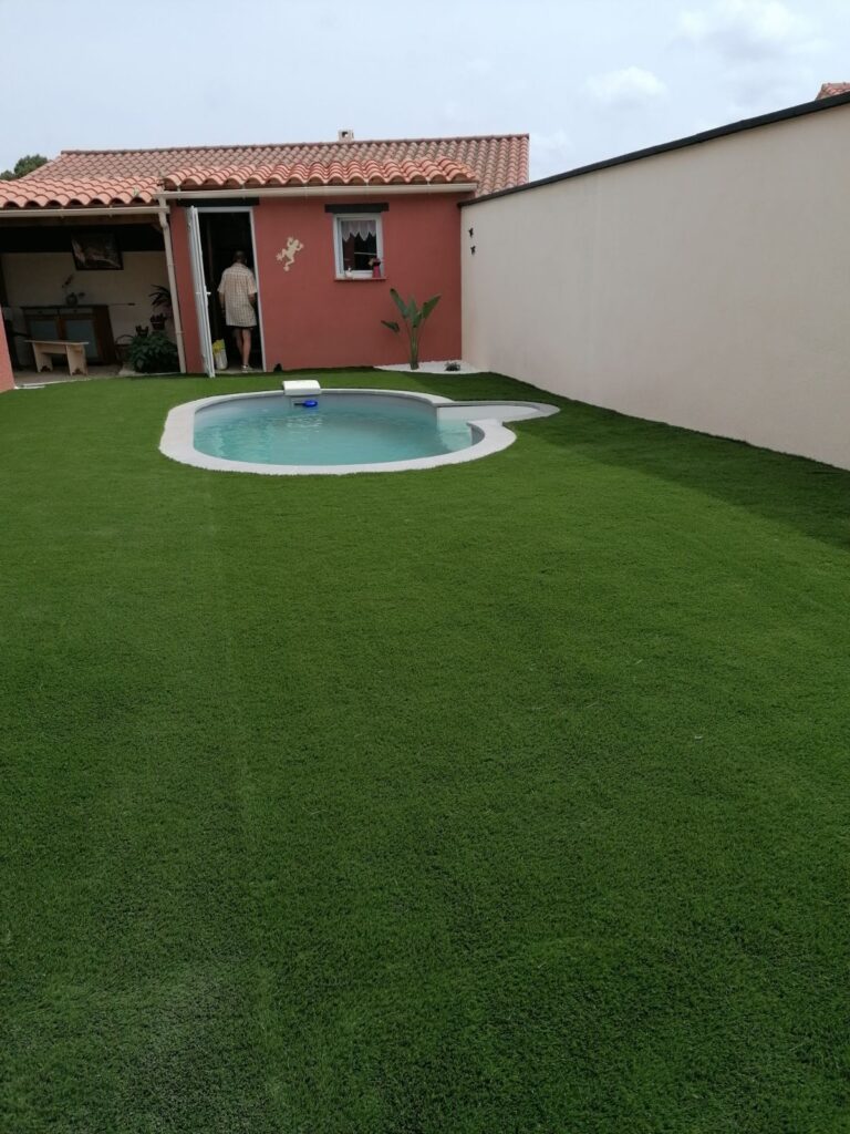Grande étendue de gazon synthétique vert encadrant une petite piscine. Annexe mur rouge, tuiles romaines et homme sortant de la porte.