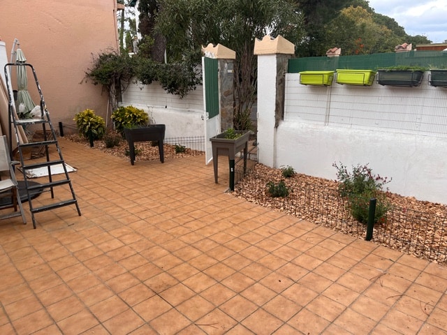 Patio extérieur avec carrelage carré terracotta. Murs blancs, parterres de gravier et plantes en pots. Une échelle de travail est posée à gauche.