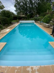 Piscine rectangulaire avec liner bleu clair, montrant les étapes immergées. Dalles beiges, jardin luxuriant, et couverture de piscine enroulée au fond.