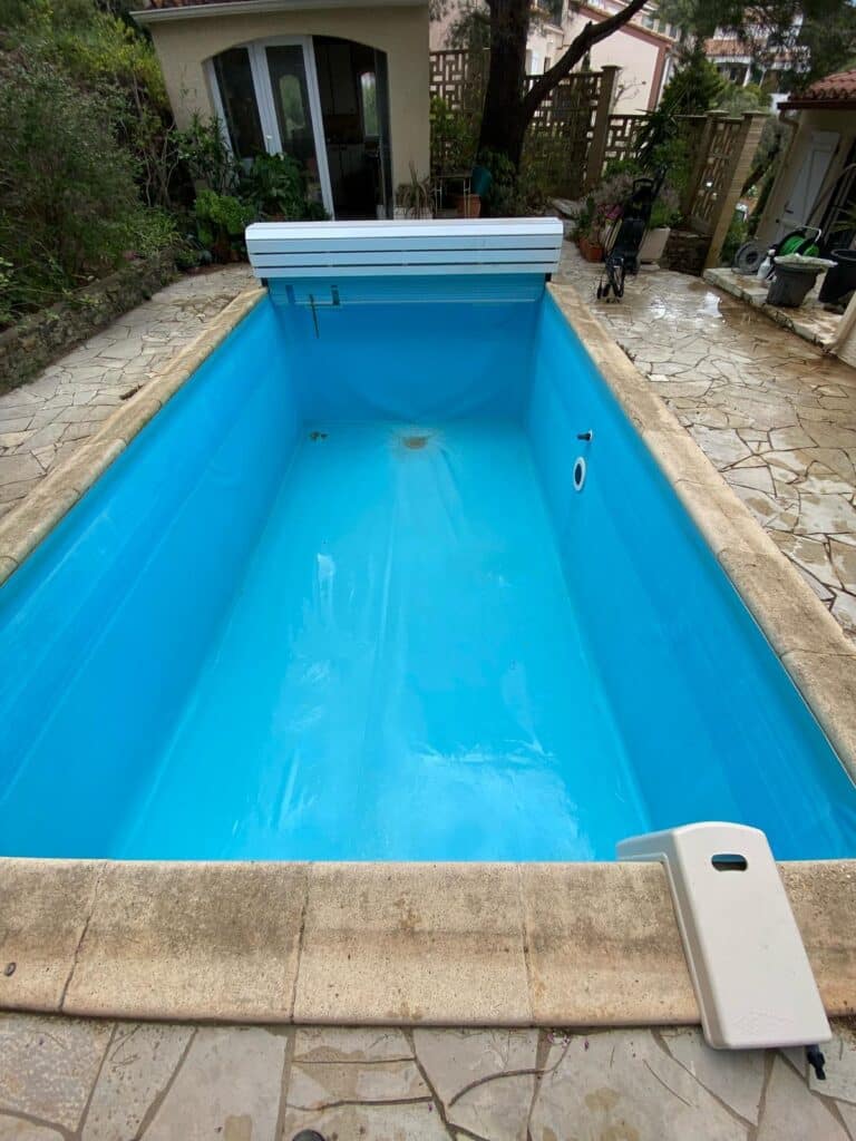 Piscine vide pour entretien : Liner bleu et couverture automatique. Piscine de jardin rectangulaire vidée avec liner bleu visible, mécanisme de volet roulant et margelles en dalles de pierre.