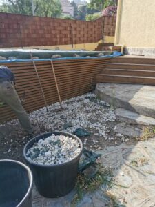 Aménagement extérieur et bassin en construction. Un ouvrier travaille près d'une paroi de lattes de bois et de marches de terrasse. Gravier blanc.